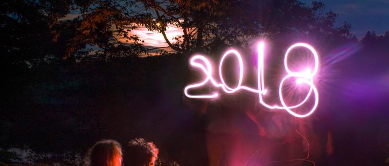 Two figures sit by the fire with the moon rising and the year 2018 written in light